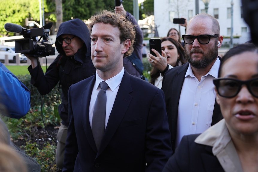 Mark Zuckerberg wearing a suit with several people walking behind and beside him.
