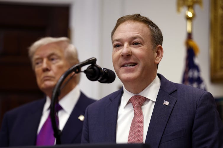 A man in a suit and tie speaks into a microphone with another man - the US President Donald Trump - behind him.