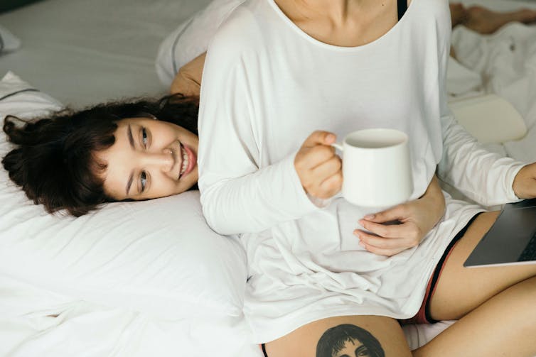 Two women snuggle on a bed while one drinks her coffee.