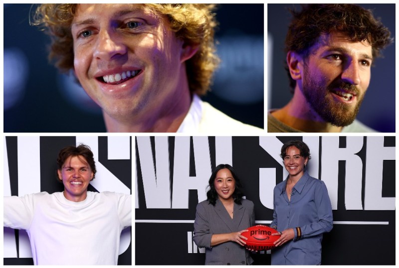 Group of smiling individuals at an event, featuring two men with curly hair and beards, along with two women holding an Australian football. The backdrop displays promotional graphics, highlighting the lively atmosphere of the occasion.