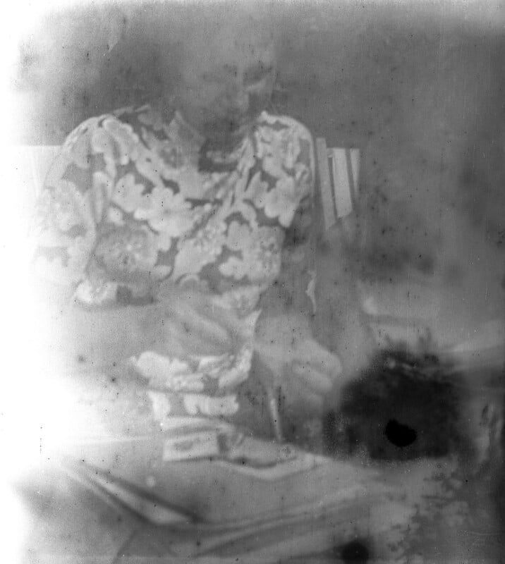A faded, blurry black and white photo of a person sitting and wearing a floral-patterned top, partially obscured by white patches and film damage. Their face and surroundings are difficult to distinguish.
