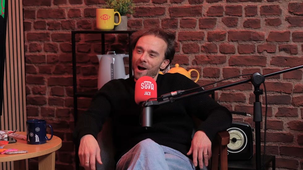 jack p shepherd on the on the sofa podcast, sitting in a chair with a microphone in front of him in a studio with a brick wall and a tea pot, kettle, and mug around him