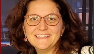 Photo of a woman with shoulder-length brown hair and glasses smiling at the camera. She appears to be indoors in front of a window at night.