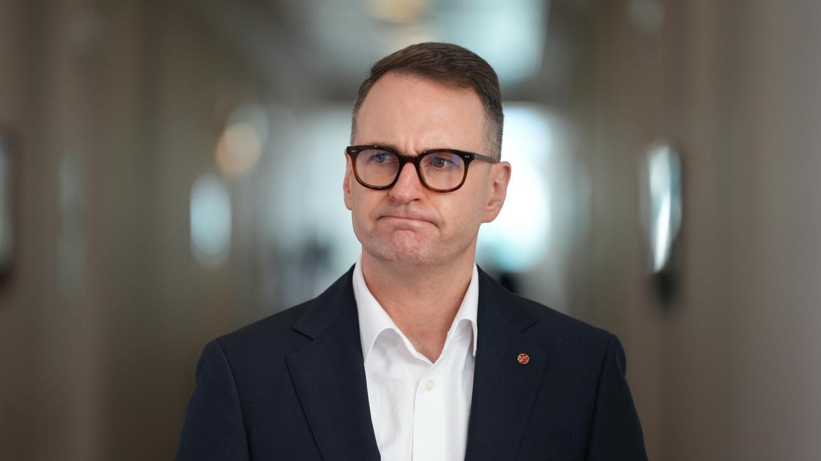 A white man wearing dark-framed glasses and a black jacket, looking frustrated.