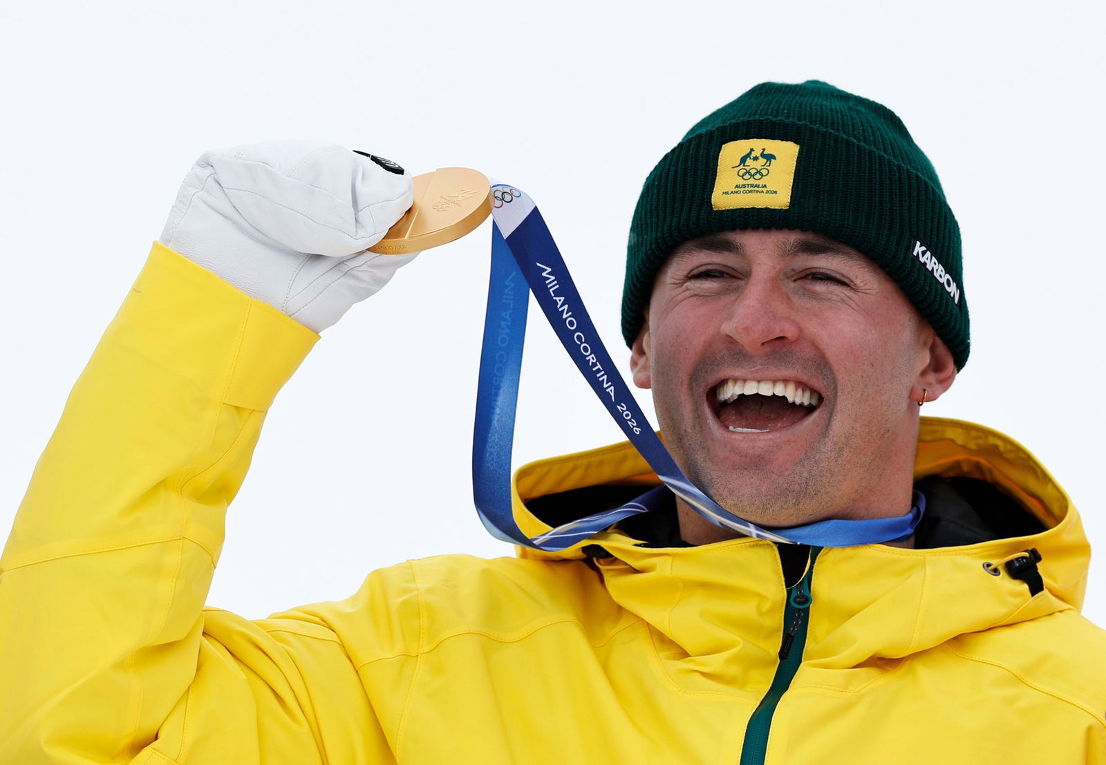 An athlete in a yellow ski jacket holds a gold medal and smiles, wearing green beanie