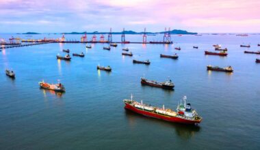 Cargo ships sit offshore near a busy port at dusk, a visual of global shipping and the climate impact of cleaner marine fuels.
