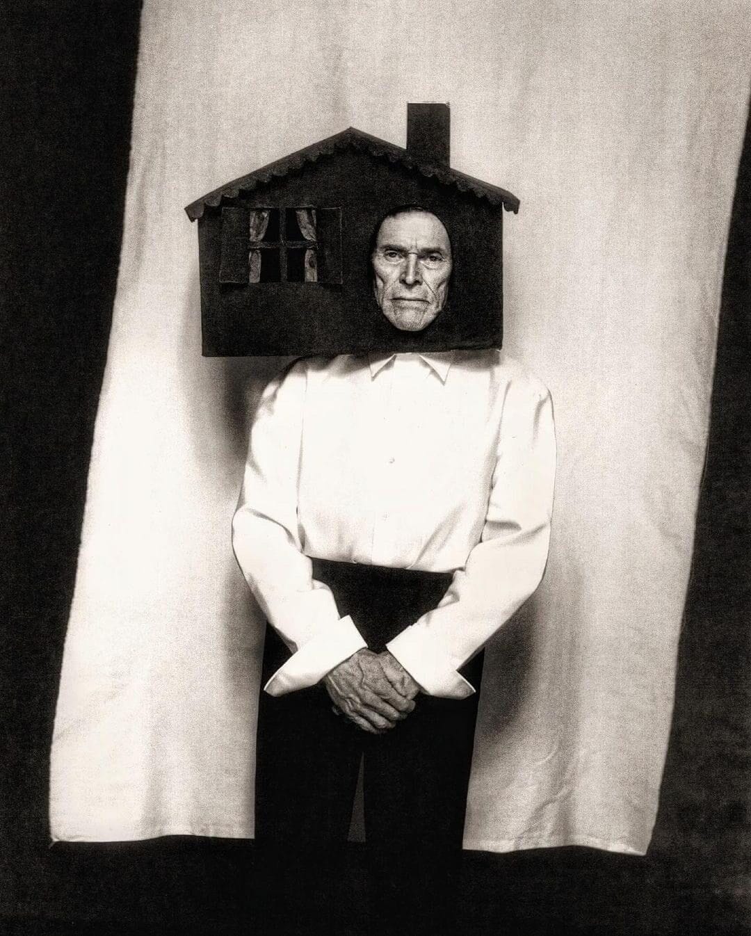 an editorial portrait by Szilveszter Makó of Willem Dafoe standing in front of a hanging white sheet, wearing a house-shaped headpiece around his face.