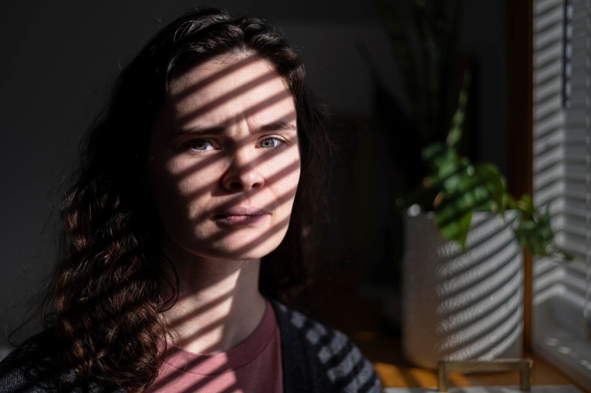 A woman with wavy brown hair stands indoors, her face partially lit by sunlight filtered through window blinds, casting shadow stripes across her face.