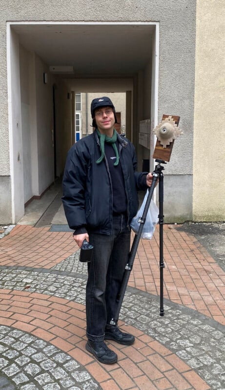 A person stands outside on a brick walkway holding a camera remote and a tripod with a handmade, wooden contraption mounted on it. The person wears a dark jacket, bucket hat, and has green headphones around their neck.
