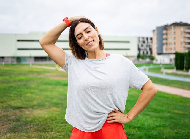 A woman between 40 and 50 years old is training outdoors in sportswear.The woman stretches her neck while standing.Concept of middle-aged women doing sports.