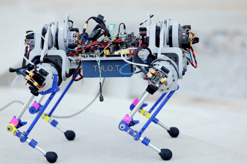 The robot has four legs with backward-facing knees. Each leg has three actuators, controlling hip ab/adduction, hip flexion/extension, and knee flexion/extension.  Exposed wires link up to the motors. T.R.O.T is emblazoned on the case around the belly of the robot.