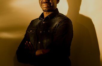 Person in dark clothing with crossed arms, casting shadow on wall: the Ugandan astronomer Geoffrey Andama.