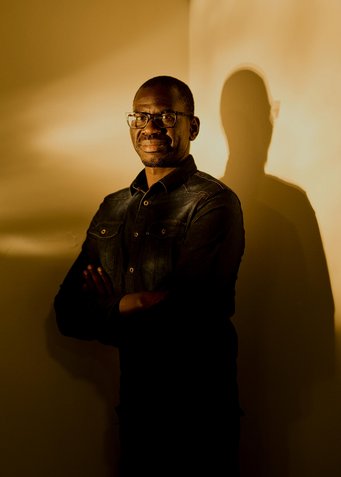 Person in dark clothing with crossed arms, casting shadow on wall: the Ugandan astronomer Geoffrey Andama.