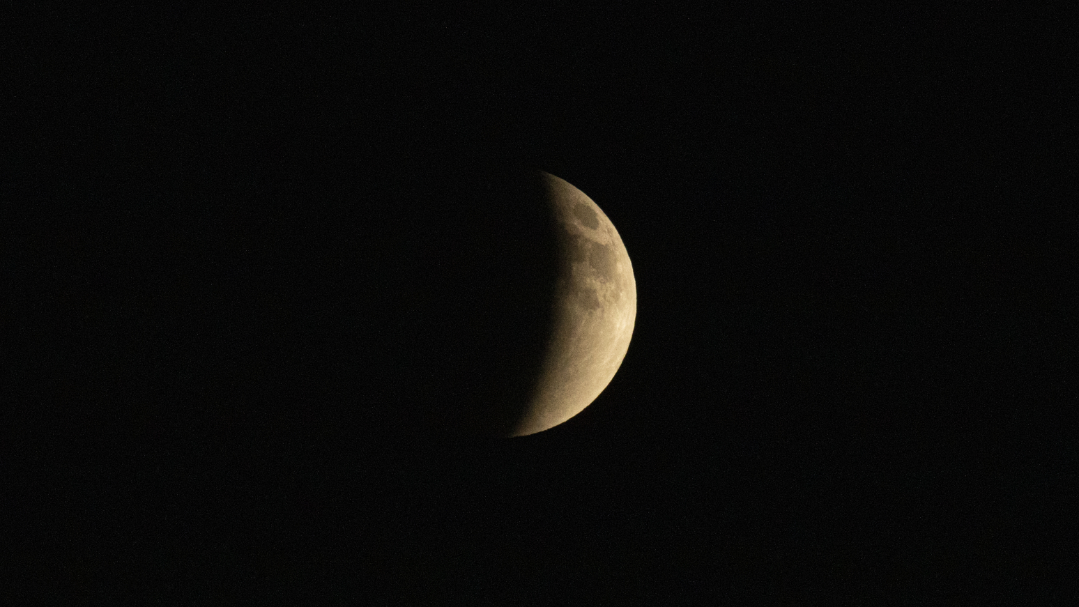 A full moon is shown during a total lunar eclipse against a black sky with its left side hidden by the curved silhouette of Earth's shadow.