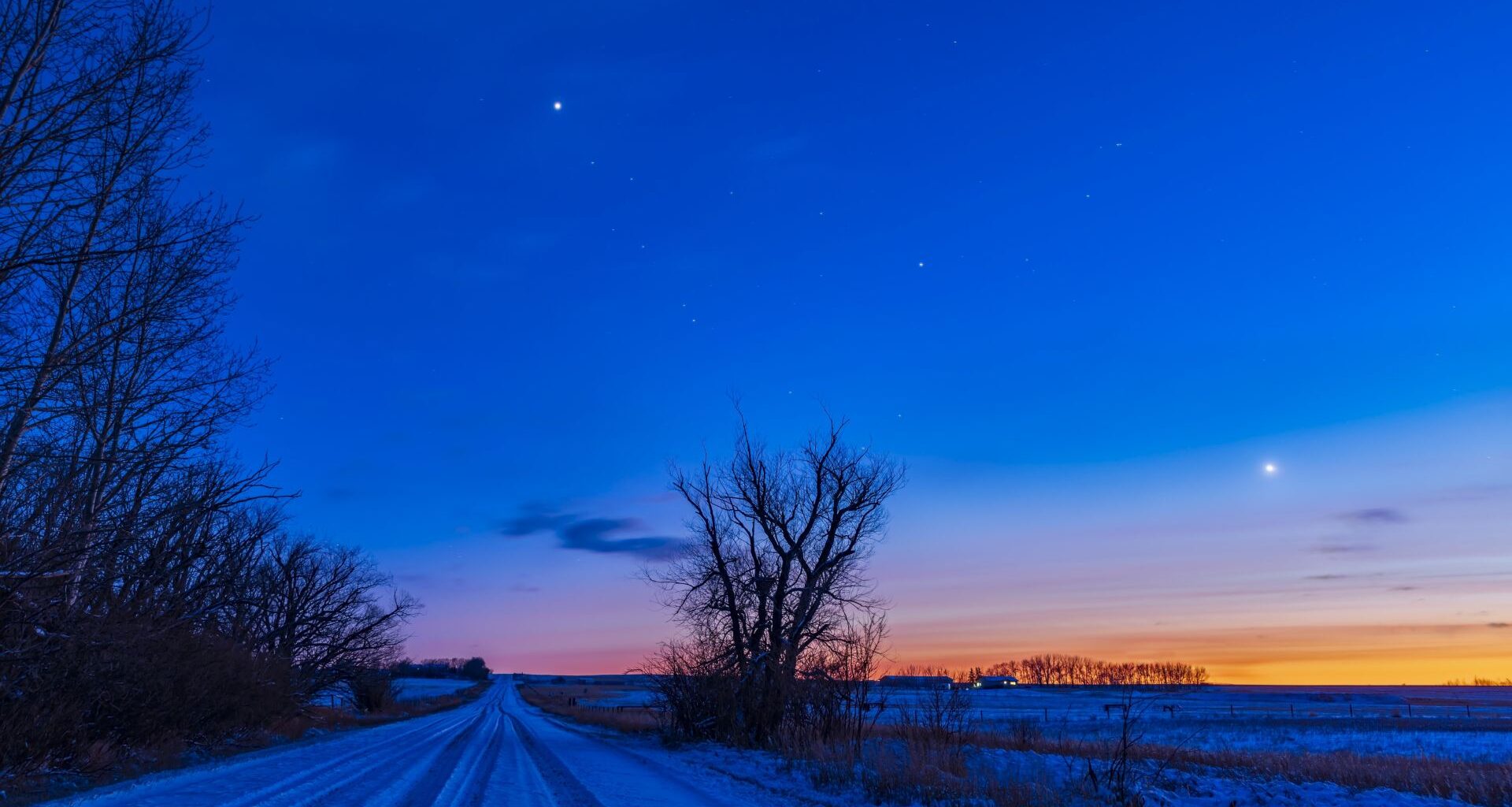 Bright star-like objects shine in the evening sky above a snowy road lined by trees on the left, as the glow of the setting sun lights up the horizon on the right of the image.
