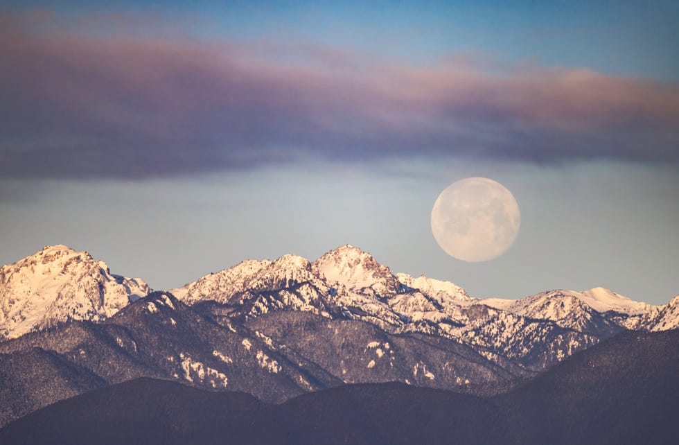 a full moon drops below a cloud bank as it sets behind snow capped mountain ridges