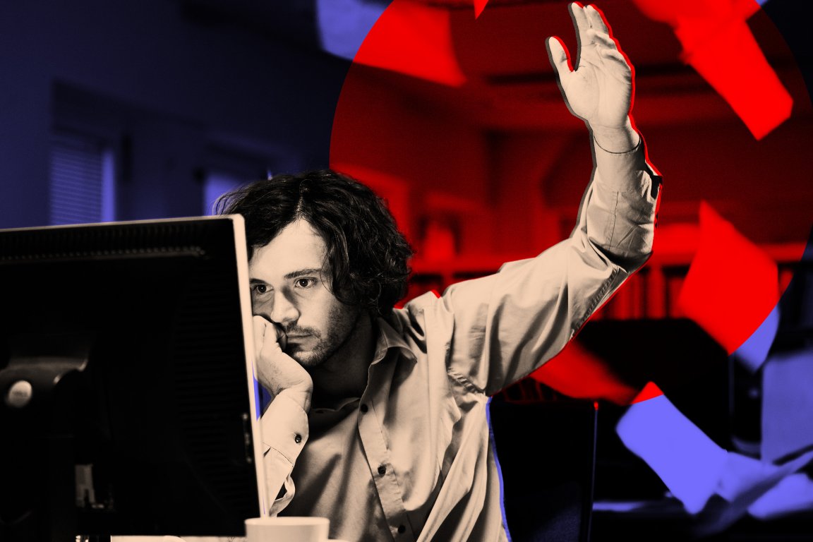 A man with curly hair and a beard sits at a desk, resting his chin on his hand while looking at a computer monitor. He raises his other hand, appearing frustrated or overwhelmed. Red and blue lighting effects highlight the background and scattered papers around him.