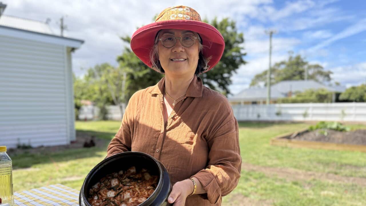 Woman in an orange hat and orange shirt smiles and holds a black pot full of kimchi