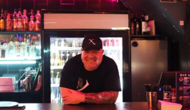 Glenn McNamara, a middle-aged white man wearing a black t-shirt and cap, leaning on a neon-lit bar.