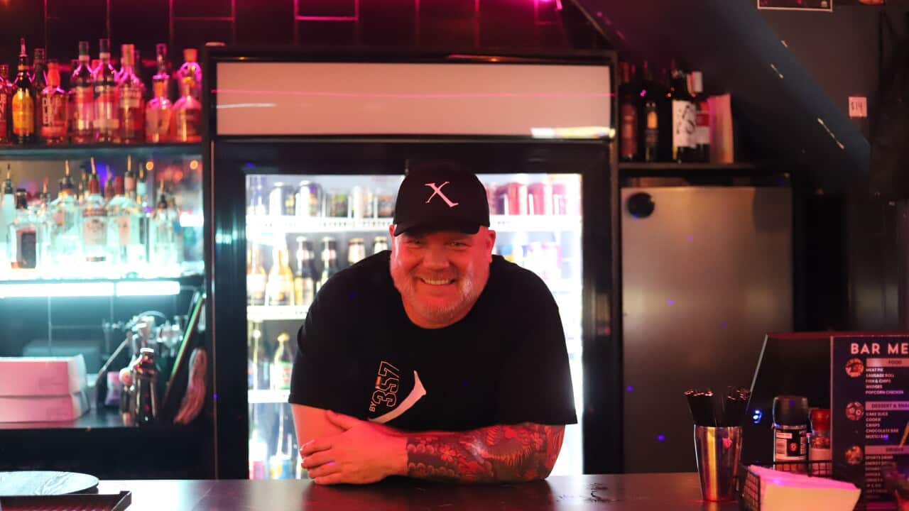 Glenn McNamara, a middle-aged white man wearing a black t-shirt and cap, leaning on a neon-lit bar.