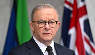 A medium shot of Anthony Albanese wearing a dark suit and glasses while standing in front of the Australian and Torres Strait Islander flags.