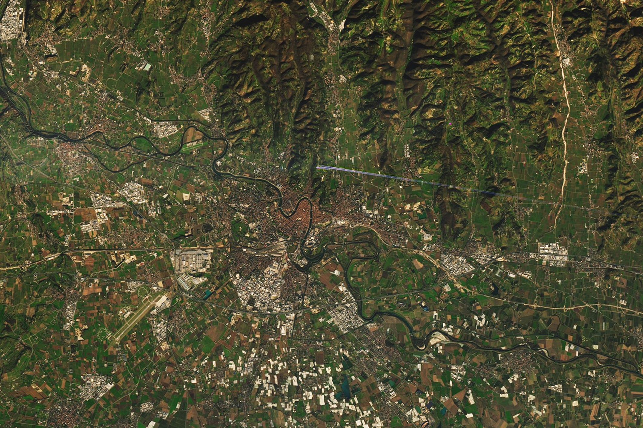 Satellite image of Verona, Italy, showing the historic city center in reddish-brown tones with dense urban development, surrounded by green agricultural fields. A river curves through the upper left, and a labeled marker highlights the ancient Roman amphitheater near the center.