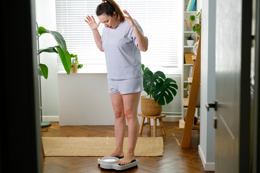 A young woman in pajamas reacts with shock and disappointment while standing on a scale at home.