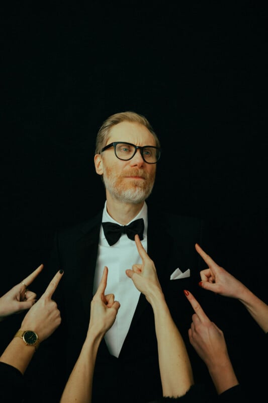 A man in a black tuxedo and glasses stands confidently against a black background while multiple hands point fingers at him from different directions.