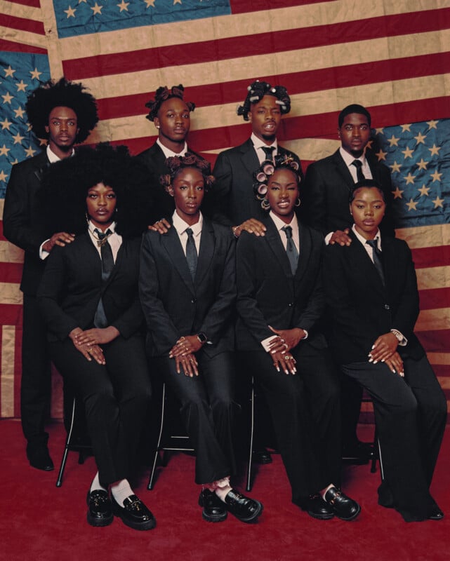 Nine Black individuals wearing black suits and ties pose formally in front of a worn American flag backdrop; some have natural hairstyles and hair rollers, and their expressions are serious.
