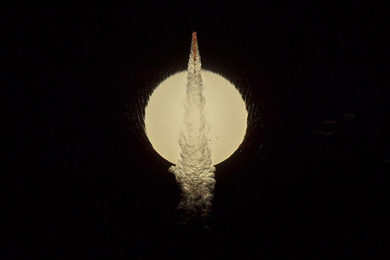 A rocket is launching directly in front of a full moon at night, with its bright exhaust plume illuminated against the dark sky and the moon’s light creating a dramatic silhouette.