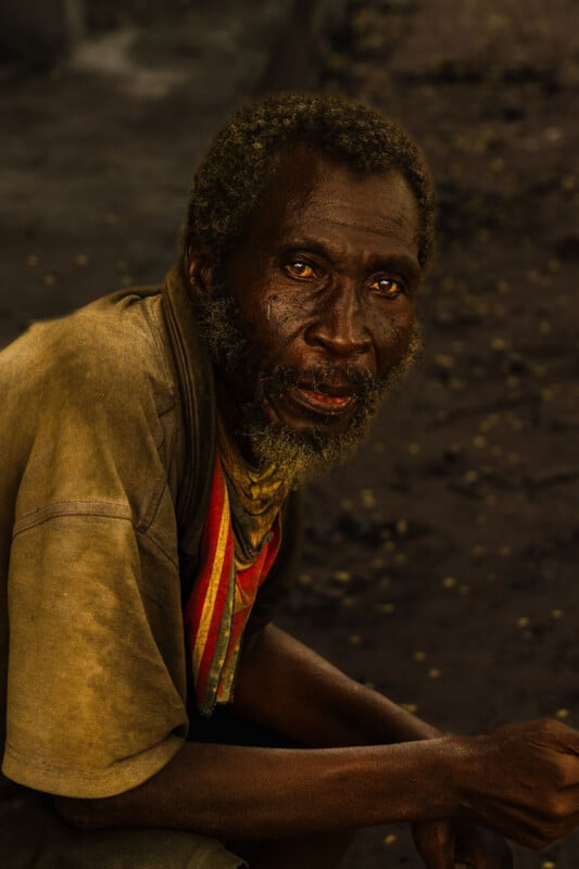A man with curly hair, a beard, and mustache sits outdoors, looking into the camera. He wears a brown shirt and a striped scarf. The background is dark and blurred, highlighting his expressive face.