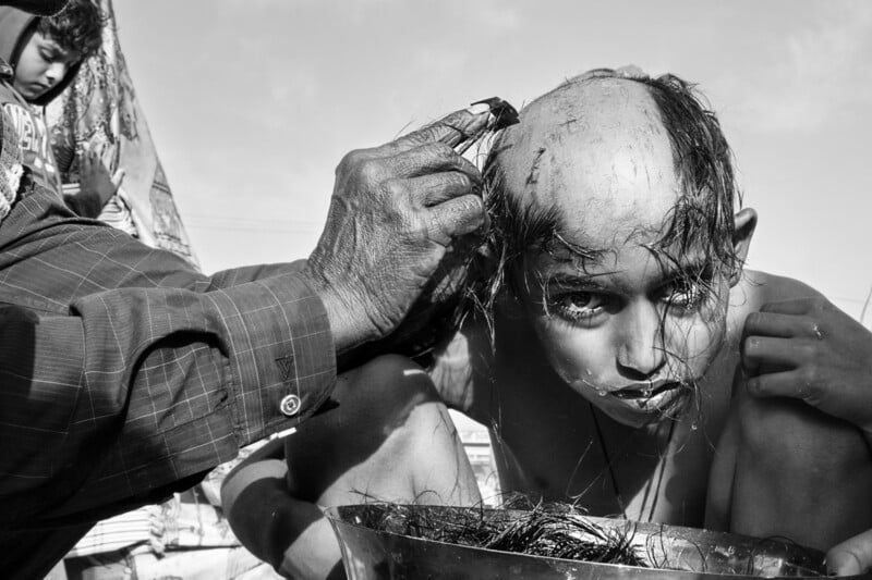 A child with a solemn expression is having their head shaved by an adult, with hair falling into a metal bowl below. The image is in black and white, highlighting the intense and focused atmosphere.