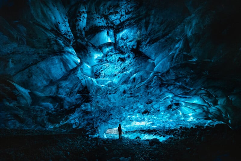 A person stands inside a vast, blue-lit ice cave, surrounded by textured, translucent ice walls and ceiling, creating a surreal and otherworldly atmosphere.