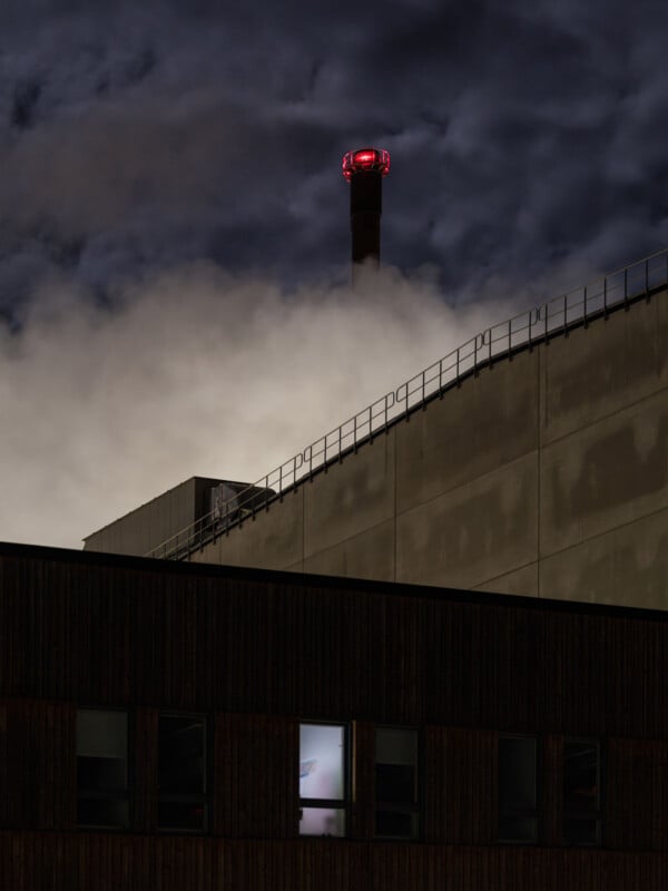 A tall smokestack with a red warning light rises above an industrial building at night, emitting wisps of smoke that blend into a dark, cloudy sky. The building has lit windows and metal railings along the roof.