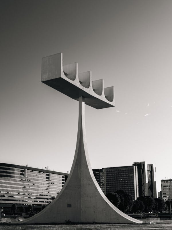 A tall concrete monument with a curved base and four rectangular prongs at the top, set against a cityscape with modern buildings in the background, photographed in black and white.