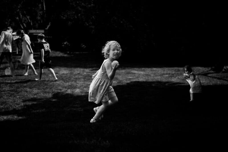 A young girl in a sunlit dress runs joyfully across a grassy area, while other children play in the background and one child stands alone to the right, all surrounded by deep shadows.