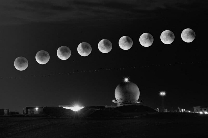 A sequence of eight phases of a lunar eclipse appears above a radar dome on a hill at night, with surrounding buildings and lights, all in black and white.