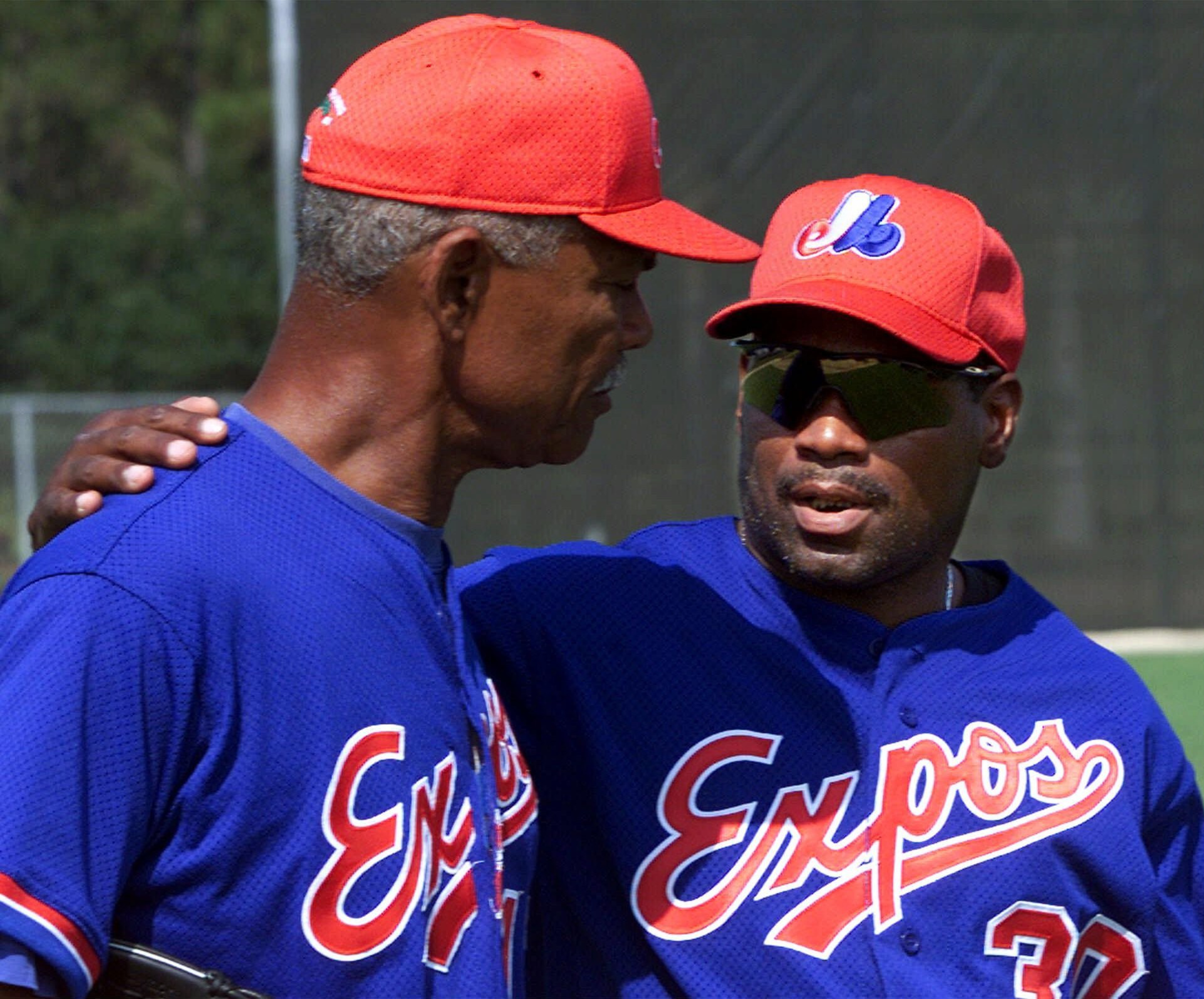 Two men in blue “Expos” baseball jerseys and red caps are standing close, with one man’s arm around the other’s shoulder, engaged in conversation.