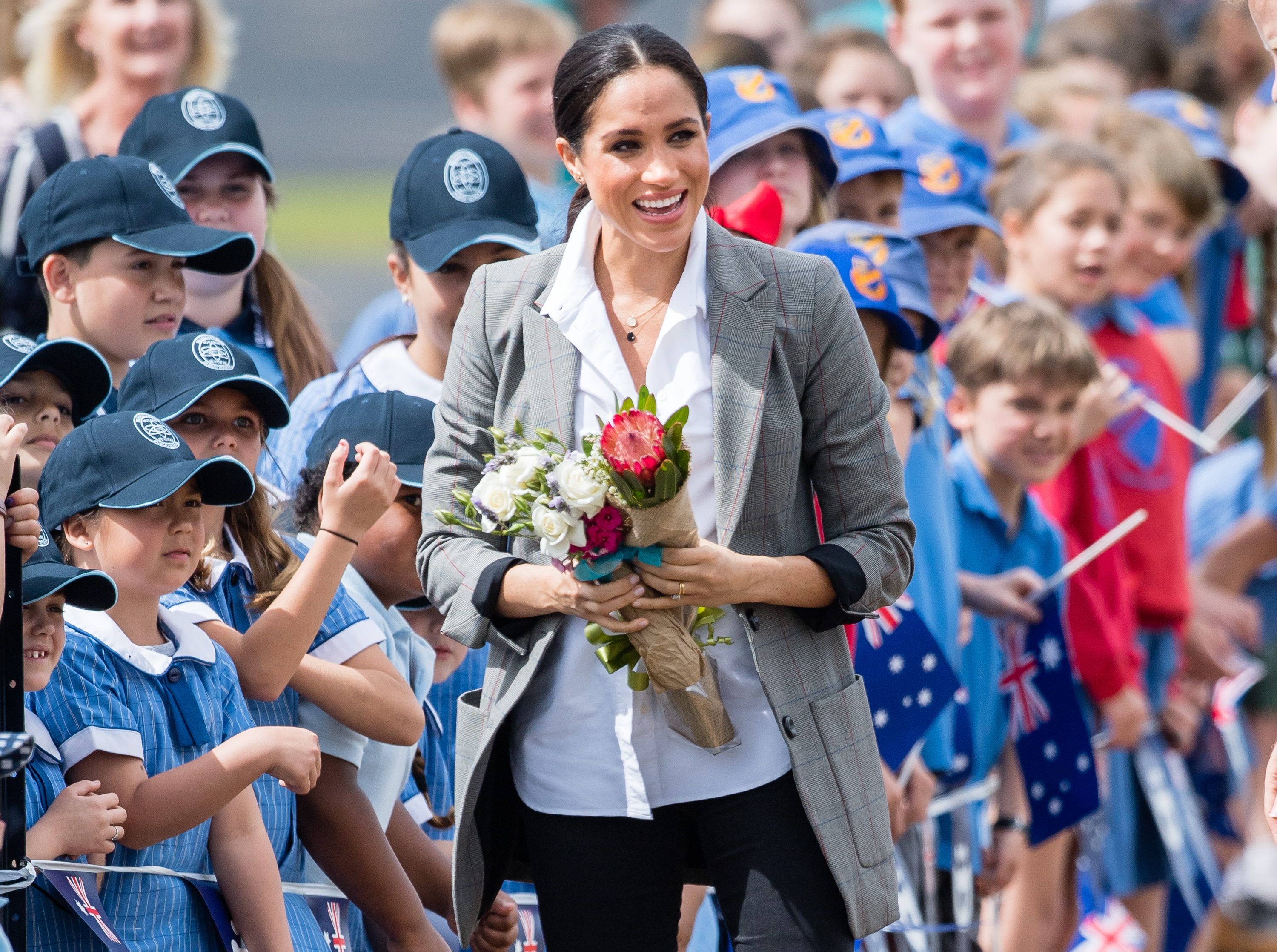 Meghan, Duchess of Sussex, smiling and holding a bouquet of flowers, surrounded by cheering children.