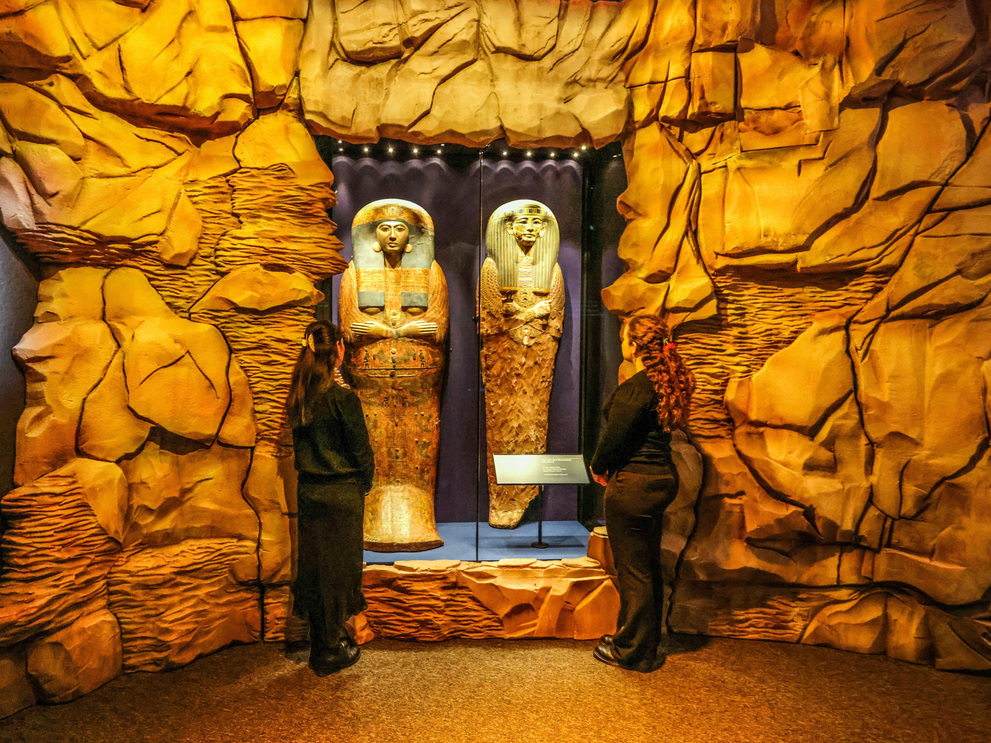 Two people facing two golden sarcophagi on display in an exhibit designed to look like a cave.