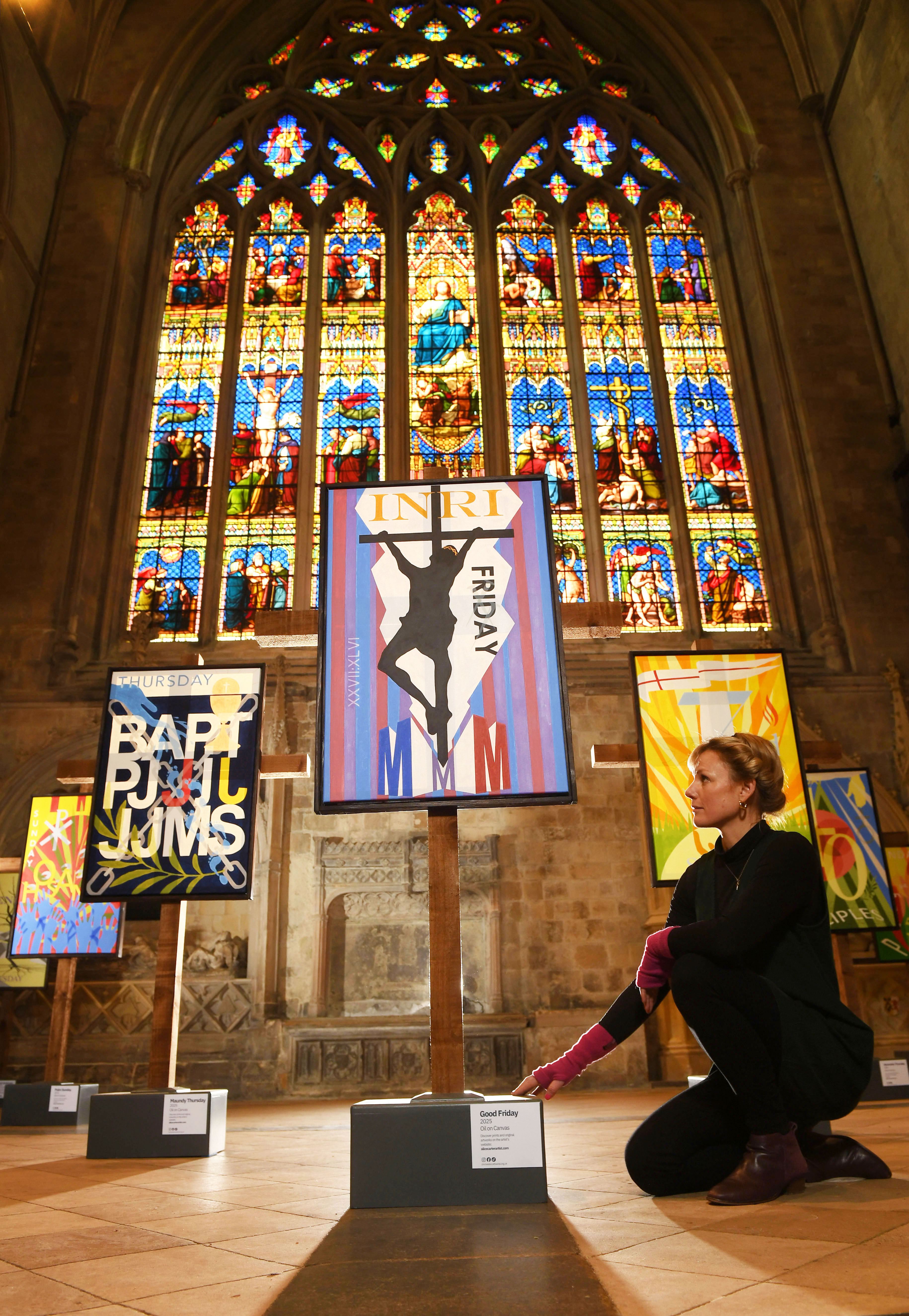Artist Alice Carter kneels beside her "Good Friday" oil painting, part of the "Ashes to Fire" series displayed in Chichester Cathedral with stained glass windows in the background.