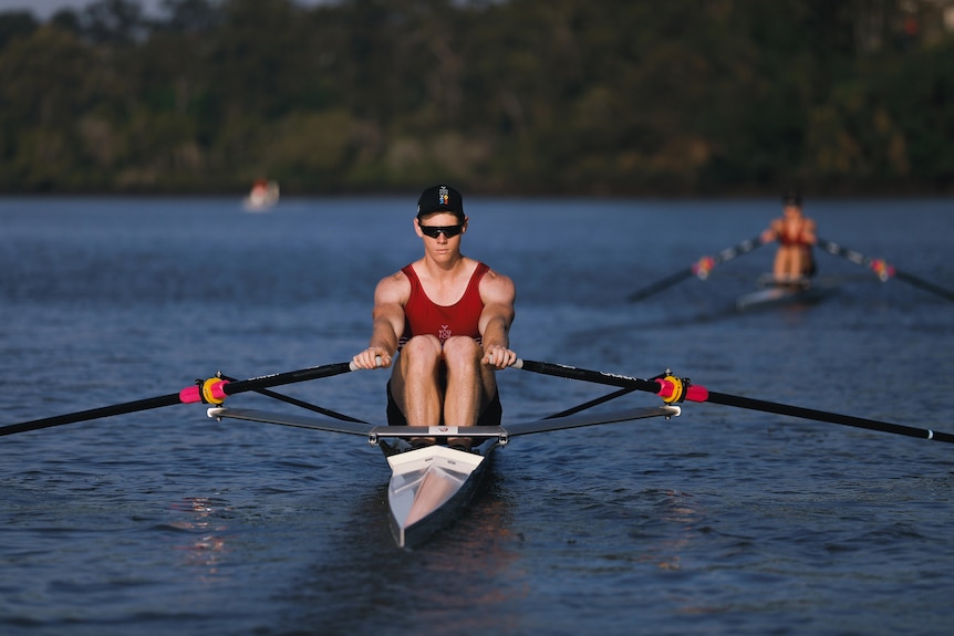 A young man rows on a river, wearing a red singlet and a black cap.