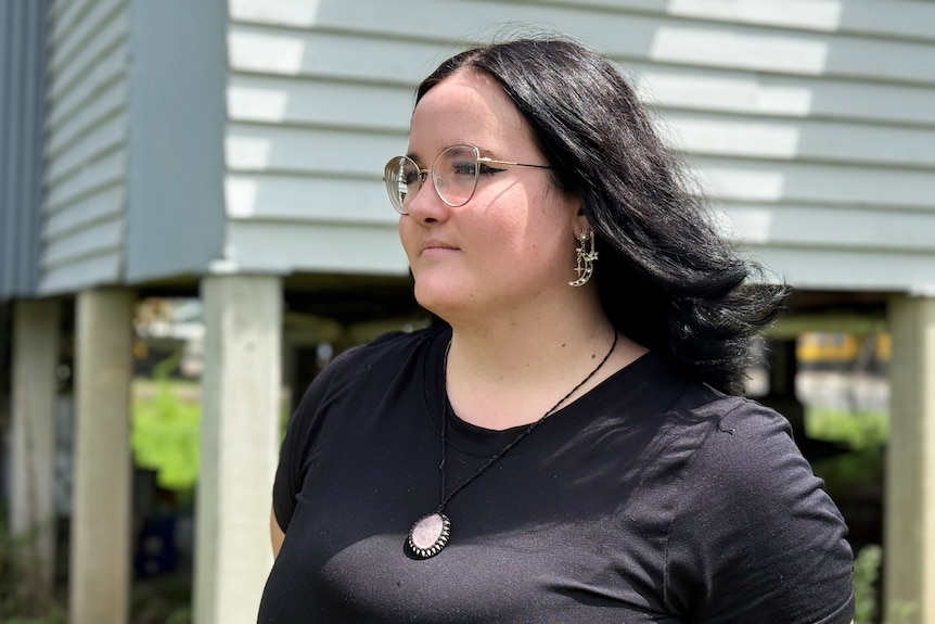 A dark-haired woman stands in front of a house on stilts.