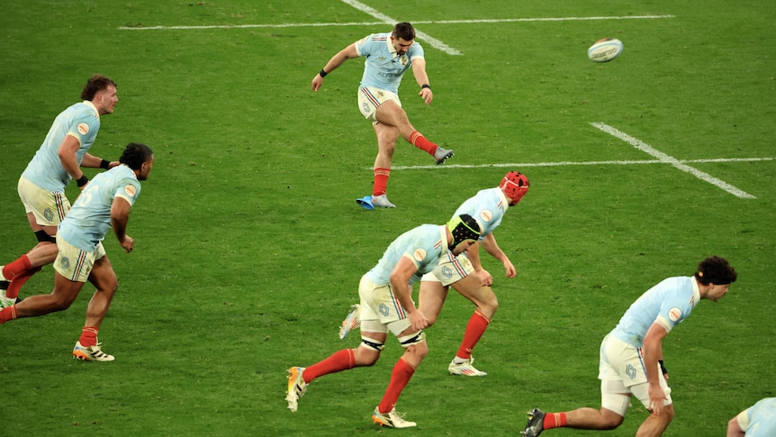 A French rugby union international kicks a ball towards the posts, as his teammates run forward.