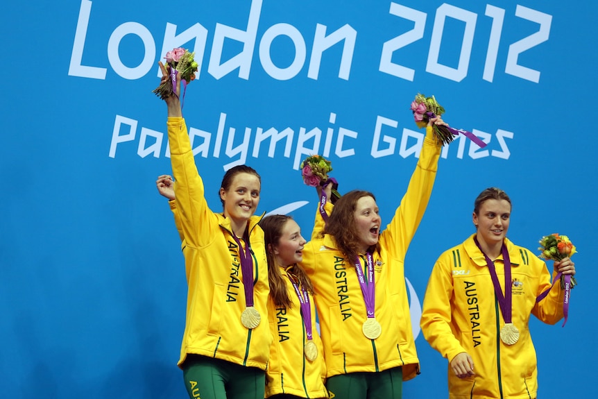 Four smiling women in Team Australia gear with gold medals around their necks hold flowers while standing on a podium.
