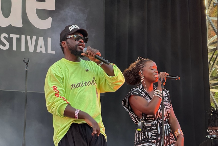 Two performers sing into microphones on a festival stage, with another musician visible in the background.