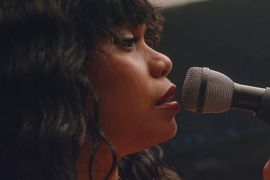 Ngaiire side profile singing into a microphone, tight head and shoulders shot.