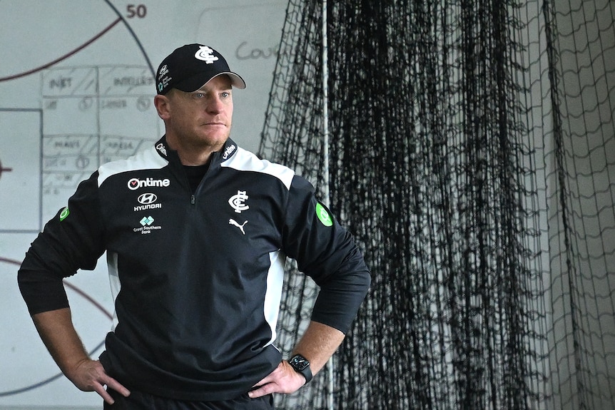 Carlton head coach Michael Voss stands with hands on hips next to nets with a whiteboard behind him.