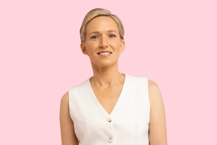 A woman with short blonde hair stands against a pink background