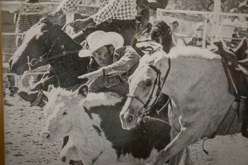 A black and white photo of a man leaning off a horse to grab a running calf